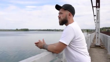 Sakallı genç bir adam köprünün yanındaki korkuluğun yanında duruyor.