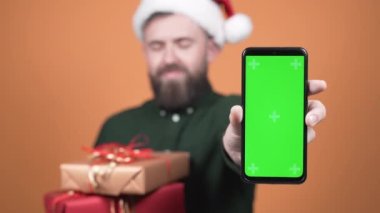 Guy in santa claus hat and gifts shows phone with green screen