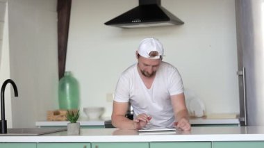 Genç adam mutfakta dururken tablete yazı yazıyor.