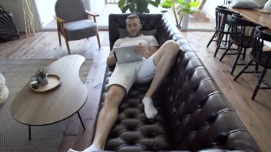 Modern apartmandaki kanepede yatarken tabletteki haberleri izleyen bir adam.