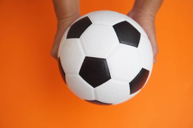 Parlak turuncu arka planda klasik bir siyah beyaz futbol topunu tutan eller, enerjiyi, takım çalışmasını ve spor ve fiziksel aktivitenin heyecanını sembolize ediyor..                               