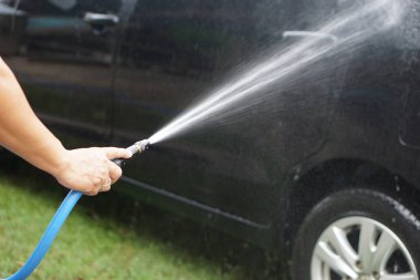 Yüksek basınçlı su hortumu kullanarak siyah bir arabayı yıkamak. Temiz su spreyi, araçların yüzeyindeki yansımalar tazelik ve temizlik gösteriyor, araba bakımını, bakımını, yaşam tarzını, temizliğini temsil ediyor.                               