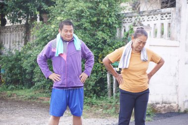 Asyalı orta yaşlı erkek ve kadın kalçalarında ellerle dışarıda ısınıyor, antrenmana hazırlanıyor, spor kıyafetleri giyiyor ve boyunlarına havlu takıyorlar. Kavram, aktif yaşlanma, refah için kişisel bakım                               