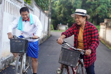 Sessiz bir mahallede Asyalı bir adamla bisiklet süren yaşlı bir Asyalı kadın, açık hava egzersizinin ve huzurlu, yeşil bir ortamda sağlıklı bir sabah rutininin tadını çıkarıyor..                               