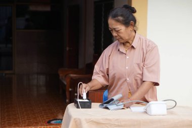 Asyalı yaşlı bir kadın, masadaki kabloları bağlayan dijital bir tansiyon monitörü hazırlıyor ve sağlığını sakin bir şekilde kontrol ediyor. Günlük sağlık kontrollerini evde yap. Sağlık hizmetleri.                                  