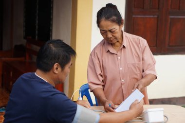 Asyalı yaşlı bir kadın evde bir erkeğin kan basıncını ölçer, sıcak aile bakımı ve dikkat gerektiren bir ev sağlığı rutini desteğini, sağlığını ve günlük şefkatini yansıtır..