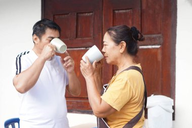   Asyalı yaşlı bir kadın orta yaşlı bir adamla açık hava ortamında sıcak bir içki içiyor. Bu an, aile bağlarını, günlük rahatlığı ve basit birlikteliği yansıtıyor..                             