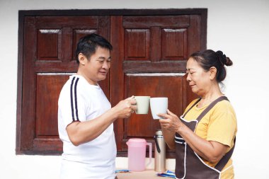 Asyalı orta yaşlı bir adam ve yaşlı bir Asyalı kadın, kahve fincanlarını dışarıda tokuşturup, basit bir evde huzurlu bir sabah ve dostça bir bağ kurarak sıcak bir anın tadını çıkarıyorlar..                               
