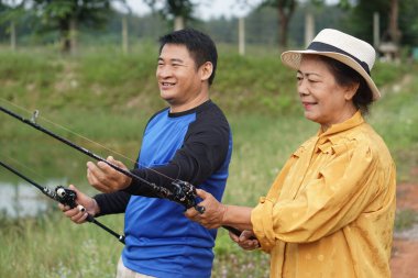       Asya 'lı son sınıf öğrencisi bir kadın ve yetişkin bir erkek, avlanmak için açık havada balık avlamaktan zevk alıyorlar. Huzurlu bir göletin yanında olta tutarken gülümsüyorlar ve doğada sıcak bir aile anını yakalıyorlar..                         