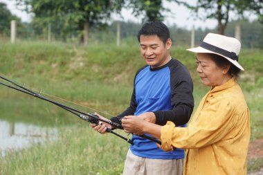       Asya 'lı son sınıf öğrencisi bir kadın ve yetişkin bir erkek, avlanmak için açık havada balık avlamaktan zevk alıyorlar. Huzurlu bir göletin yanında olta tutarken gülümsüyorlar ve doğada sıcak bir aile anını yakalıyorlar..                         