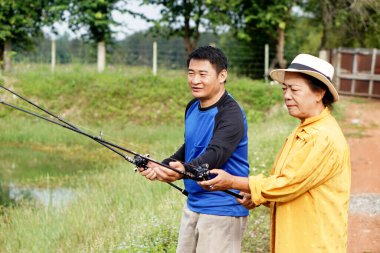       Asya 'lı son sınıf öğrencisi bir kadın ve yetişkin bir erkek, avlanmak için açık havada balık avlamaktan zevk alıyorlar. Huzurlu bir göletin yanında olta tutarken gülümsüyorlar ve doğada sıcak bir aile anını yakalıyorlar..                         