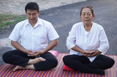 Orta yaşlı bir Asyalı erkek ve kadın açık hava minderlerinde bacak bacak üstüne atarak birlikte huzurlu meditasyon yapıyorlar. Her ikisi de basit beyaz elbiseler giyiyor, gözleri kapalı, sakin bir farkındalık ve rahatlama atmosferi yaratıyor..                               