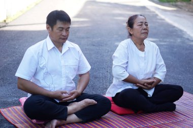 Orta yaşlı bir Asyalı erkek ve kadın açık hava minderlerinde bacak bacak üstüne atarak birlikte huzurlu meditasyon yapıyorlar. Her ikisi de basit beyaz elbiseler giyiyor, gözleri kapalı, sakin bir farkındalık ve rahatlama atmosferi yaratıyor..                               