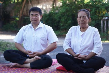 Orta yaşlı bir Asyalı erkek ve kadın açık hava minderlerinde bacak bacak üstüne atarak birlikte huzurlu meditasyon yapıyorlar. Her ikisi de basit beyaz elbiseler giyiyor, gözleri kapalı, sakin bir farkındalık ve rahatlama atmosferi yaratıyor..                               