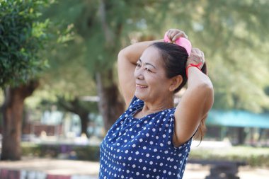 Mutlu son sınıf Asyalı kadın saçını dışarıda parkta topluyor. Kendine bakmaktan, sağlıklı yaşam tarzından ve doğal güzellikten hoşlanan kendine güvenen bir kadın, kadınların hiçbir yaşta güzel olmayı bırakmadığını gösterir..