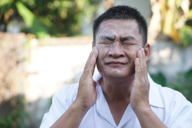 Middle aged Asian man applying facial cream outdoors, eyes closed, gently massaging cheeks for skincare routine. Concept of healthy skin, men grooming, anti aging, self care, and daily facial care.