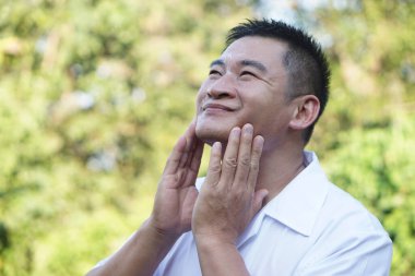 Orta yaşlı Asyalı adam dışarıda yüz kremi sürüyor, gözleri kapalı, cilt bakımı için yanaklarına nazikçe masaj yapıyor. Sağlıklı cilt, bakımlı erkekler, yaşlanma karşıtı, kişisel bakım ve günlük yüz bakımı kavramı.