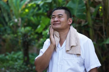 Orta yaşlı Asyalı bir adam açık havada havluyla yüzüne dokunuyor, tımar sonrası temiz havanın tadını çıkarıyor. Erkekler cilt bakımı ve kişisel hijyen konsepti, sağlıklı yaşam tarzı, kişisel bakım, güven ve rahatlama.