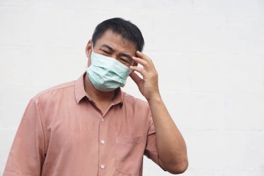 Asyalı adam tıbbi maske takıyor. Şakağına yorgun bir ifadeyle dokunuyor. Baş ağrısı, stres ya da hastalık hissediyor. Sağlık sorunu kavramı, yorgunluk, virüs koruması, salgın yaşam tarzı, zihinsel gerginlik