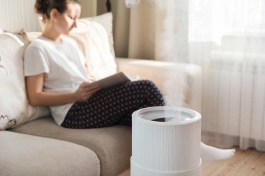 Modern hava nemlendirici, evde aroma yağı yayıcı. Bir evde yaşamanın rahatlığını geliştirmek, refahı arttırmak. Ultrasonik buhar teknolojisi. Arka planda kitap okuyan kadın
