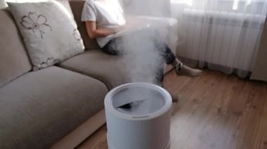 Modern hava nemlendirici, evde aroma yağı yayıcı. Bir evde yaşamanın rahatlığını geliştirmek, refahı arttırmak. Ultrasonik buhar teknolojisi. Arka planda kitap okuyan kadın