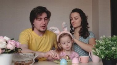 Mutlu Paskalyalar. Aile Paskalya için hazırlanıyor. Çocuklu ebeveynler Paskalya yumurtalarını boyar. Anne, baba ve kız tavşan kulağı takıyor. Gülen yüzler.