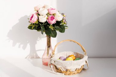Beyaz ahşap masada, sepetteki Paskalya renkli yumurtalar. Arka planda bir buket gül çiçeği. Mutlu Paskalyalar. İlkbahar havası.
