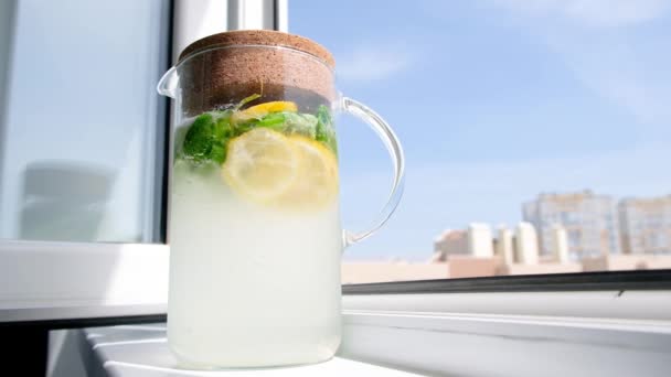Домашній лимонад у скляному глечику на вікні. Літній освіжаючий напій. Холодна детоксикаційна вода з лимоном — стокове відео