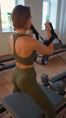 A woman in activewear does pilates exercises for the chest, arms and back muscles with a reformer machine in a sunlite studio, committed to her workout routine