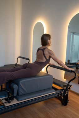 Aktif giyimli bir kadın güneşli bir stüdyoda göğüs ve sırt kasları için egzersiz yapıyor. Egzersiz yapmaya kendini adamış.