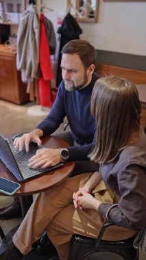 İş arkadaşları proje planlıyorlar ve kafeterya masasında büyük pencereli evrakları gözden geçiriyorlar. Proje planlaması, evrak işleri, kafe masası, takım çalışması, tartışma.