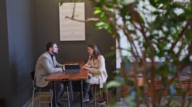 İş arkadaşları proje planlıyorlar ve kafeterya masasında büyük pencereli evrakları gözden geçiriyorlar. Proje planlaması, evrak işleri, kafe masası, takım çalışması, tartışma.