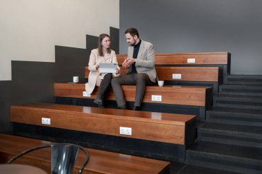 Modern çalışma alanındaki katmanlı ahşap koltuklarda iş ortakları sohbet ediyor. Konuşma, sıralı oturma, takım çalışması, çalışma alanı, modern ofis.