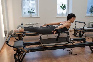 Pilates reformatöründe yan kalas tutan bir kadın. Spor salonunda bacaklarını açmış, pencere pencere gezen bir kadın. Yan tahtası, pilates reformatörü, bacakları açılmış, stüdyo, pencere..