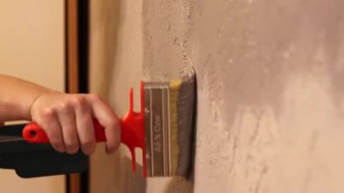 Kapalı alanda duvar boyamak, seçici bir odak noktası. Geniş bir fırçayla duvarı yeniden boyamak. Tamir ve boyama işi. Bir kadının eline bir fırçayla yakından bak. Duvarı gri boyayla boyarken.