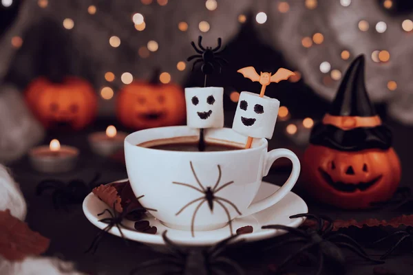 Cadılar Bayramı kahvesi. Arka planda çubukların, örümceklerin ve balkabaklarının üzerinde iki tane gülen marşmelov hayaleti. Sıcak, neşeli ve ürkütücü sonbahar içkisi sahnesi..