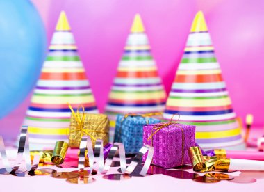 Renkli parti şapkaları, küçük hediye kutuları, konfeti ve parlak pembe arka plandaki flamalar şenlikli bir kutlama sahnesi oluşturur. Yakın plan..