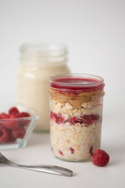 Gece yulafları ahududu dolu cam bir kavanozda, merkezde, yan görüş, seçici odak, beyaz arka plan ve masa, parlak ve havadar görünüm