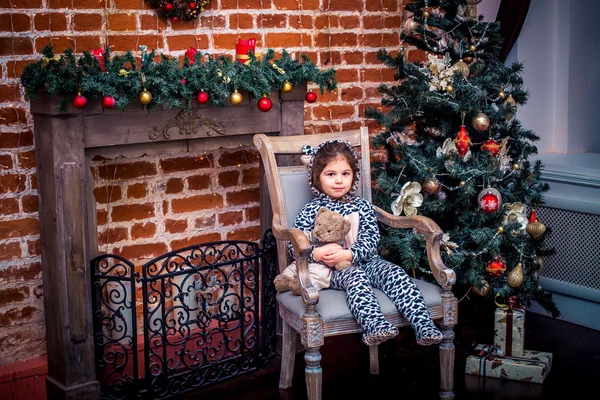 Szép kislány, mosolygós, mackó közelében a karácsonyfa, vintage széken ül. Boldog Új Évet. — Stock Fotó