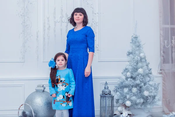 Happy mother with her daughter in long blue dresses  stand near the Christmas tree