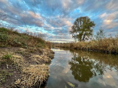 Nehir yavaşça aşağı akıyor, günbatımı bulutlarını yansıtıyor ve kuru bitki örtüsünü kuşatıyor.