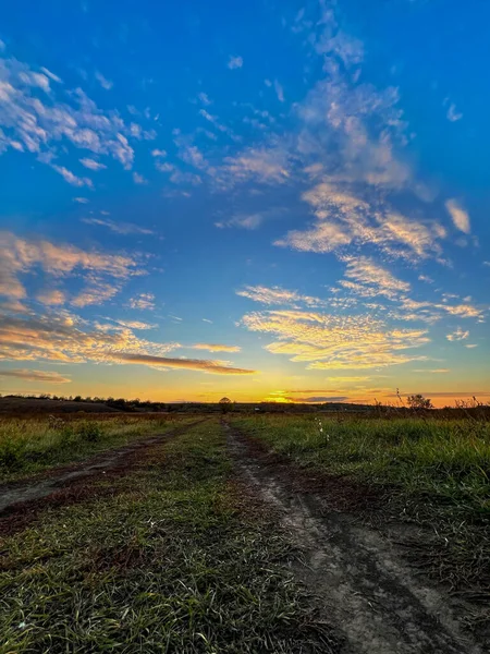 Toprak yol, bulutlarla dolu, canlı bir günbatımı gökyüzünün altında yeşil bir tarladan geçiyor.
