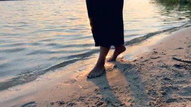Sıcak, altın bir günbatımında, kıyı şeridi boyunca çıplak ayakla yürüyen bir kadın.