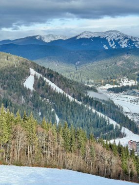 Kış manzarası kar kaplı tepeleri olan yeşil ağaçların arasından süzülen kayak yamaçlarını gösteriyor.