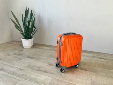 Turuncu renkli tekerlekli bavul, köşesinde bir bitkiyle birlikte açık ahşap zeminde duruyor.