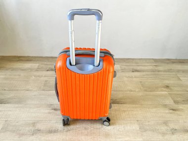 Teleskop sapı ve tekerlekleri olan turuncu kabuklu bavul ahşap bir zeminde duruyor, yolculuğa hazır.