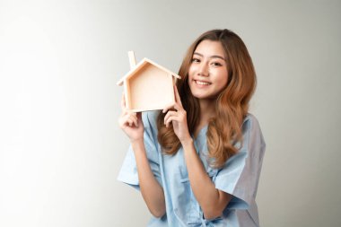 Ahşap ev modelini tutan genç ve güzel Asyalı bir kadının portresi. Fotokopi alanı, kendini karantina, sağlık sigortası programı olan evde kendini mutlu ve mutlu hissediyor.