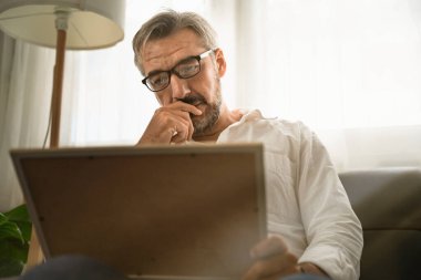 Üzgün, yaşlı bir adam kanepeye oturmuş resim çerçevesine bakıyor ve resimdeki birini düşünüyor. Covid-19 salgınından sonra ölen birinin kaybolması kavramı.