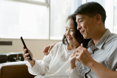 Sahnedeki yaşlı Asyalı çift, evde tek başına dolaşırken akıllı telefondan bir video konferans görüşmesi yapıyor..