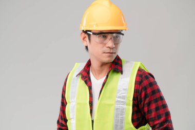 Sarı şapka takan genç Asyalı ustabaşı portresi ya da gri arka planda gözlük koruması ve güvenlik yeleği takan güvenlik kaskı. Güvenlik üniforması portresi kavramı.